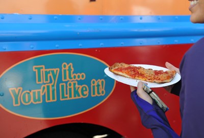 A person holding a plate with a slice of pizza, in front of a sign that reads "Try it...You'll like it!"